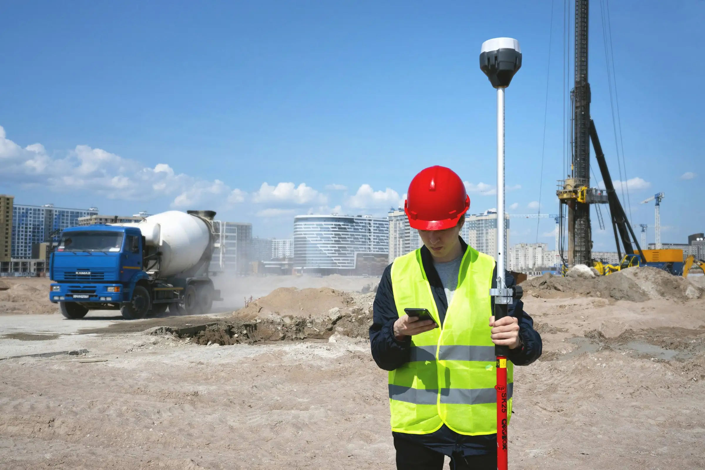 Site engineer in a hard hat checking a phone beside a survey pole on an active construction site