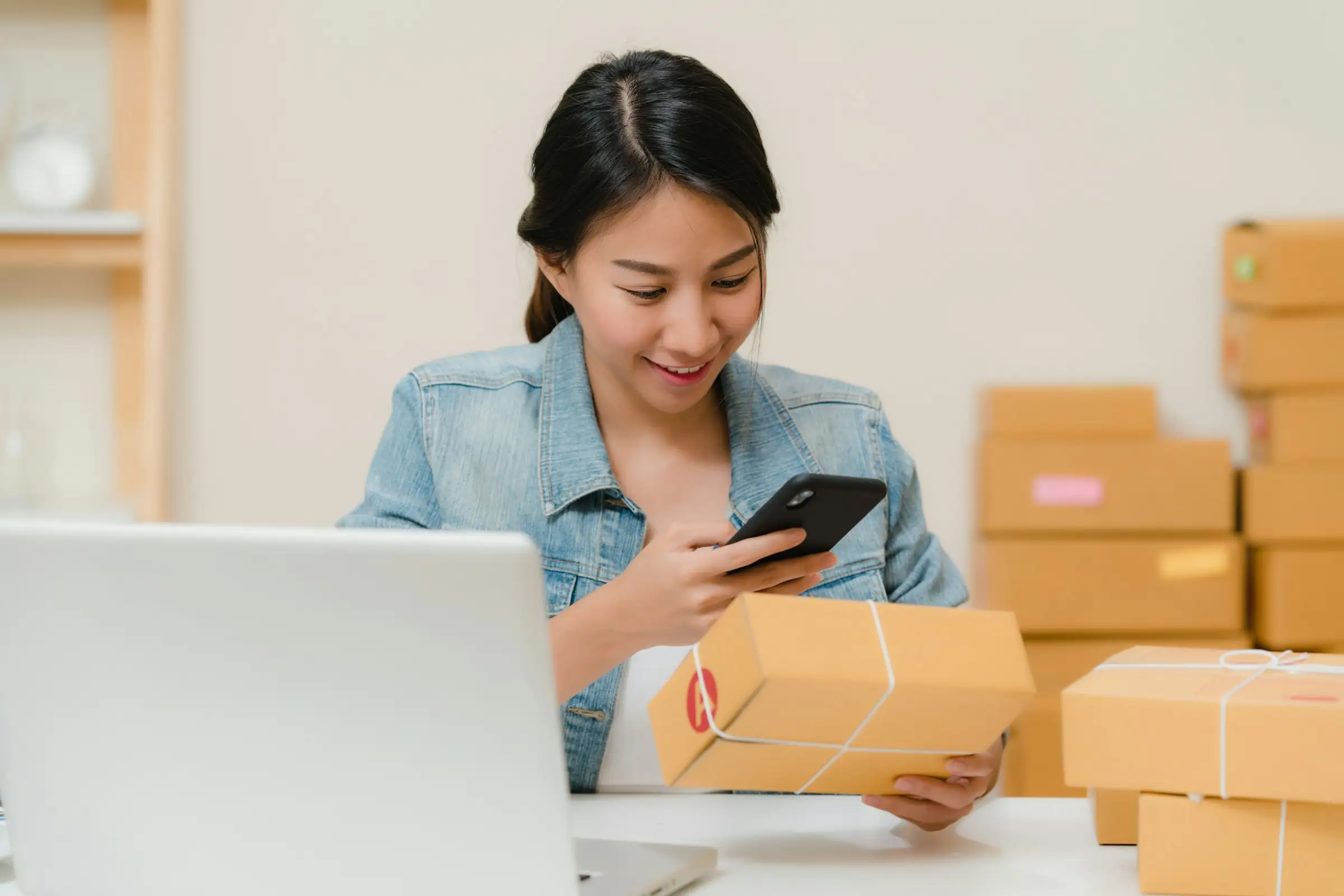 Online seller scanning a shipping package with her phone beside a laptop and boxes