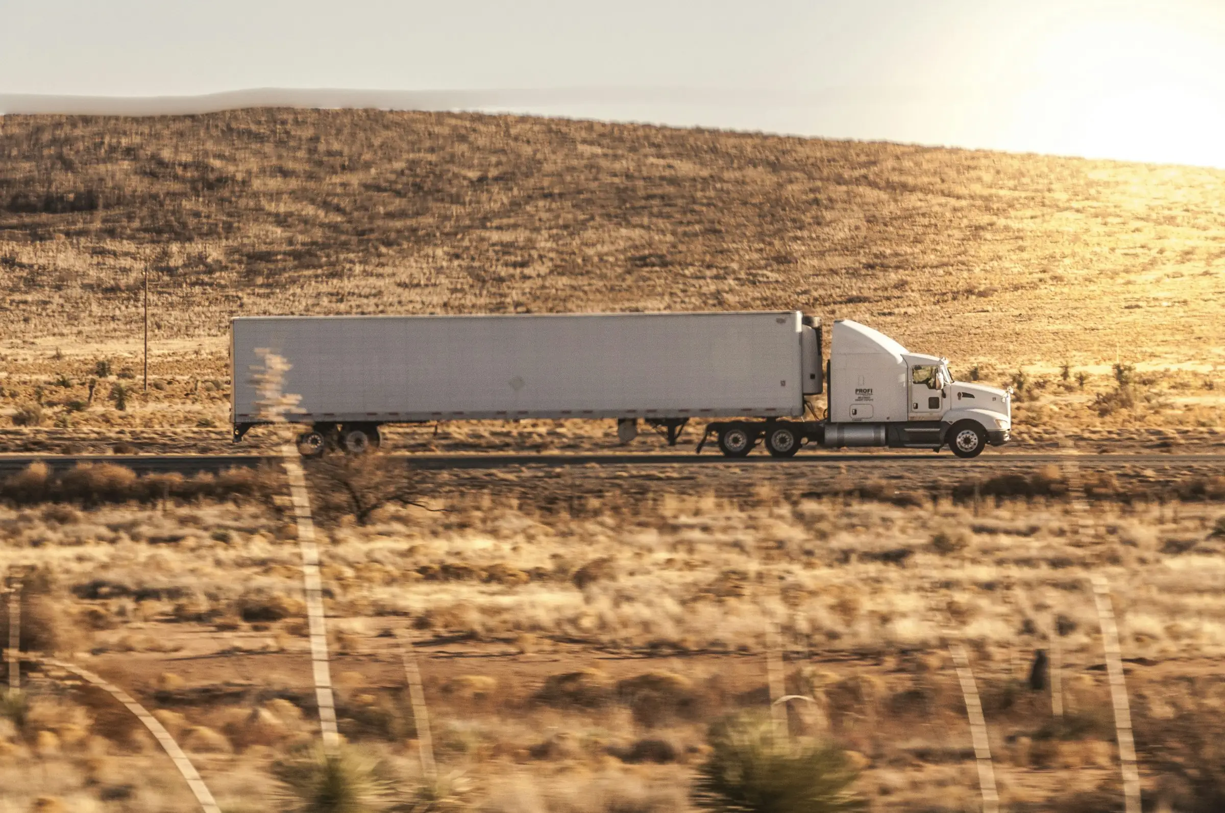 White semi-truck hauling a trailer along a rural highway at golden hour